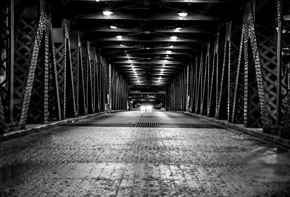 A black and white image of an industrial steel bridge, lit by overhead lights. The perspective shows the bridge's interior structure with cross-braced beams along the sides. In the distance, headlights of an approaching vehicle are visible, creating a sense of depth and symmetry.
